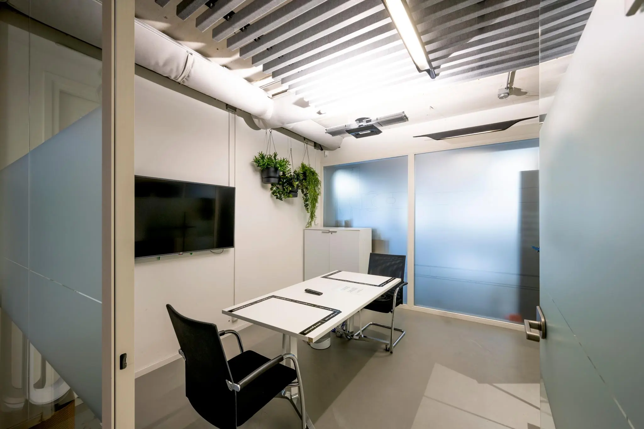 Moderne vergaderruimte aan de Fred. Roeskestraat met glazen wanden, een tafel met twee stoelen, een flatscreen en hangende planten.